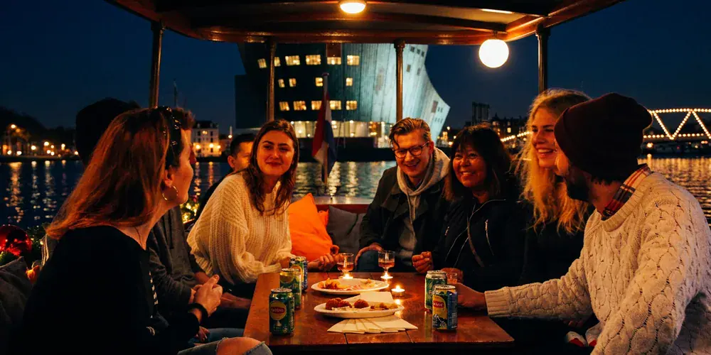 Groep borrelt 's avonds met bitterballen in een salonboot tijdens het Amsterdam Light Festival